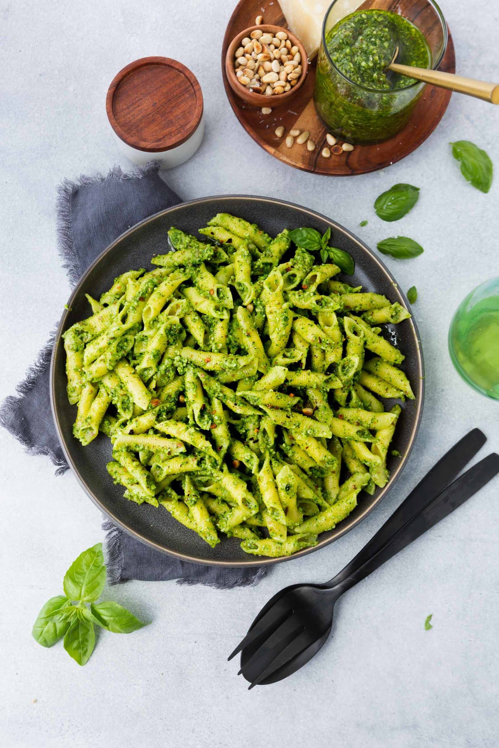 a photo of a plate of cooked penne pasta covered in bright green pesto with a cup of fresh pesto next to it and a jar of pine nuts on the side