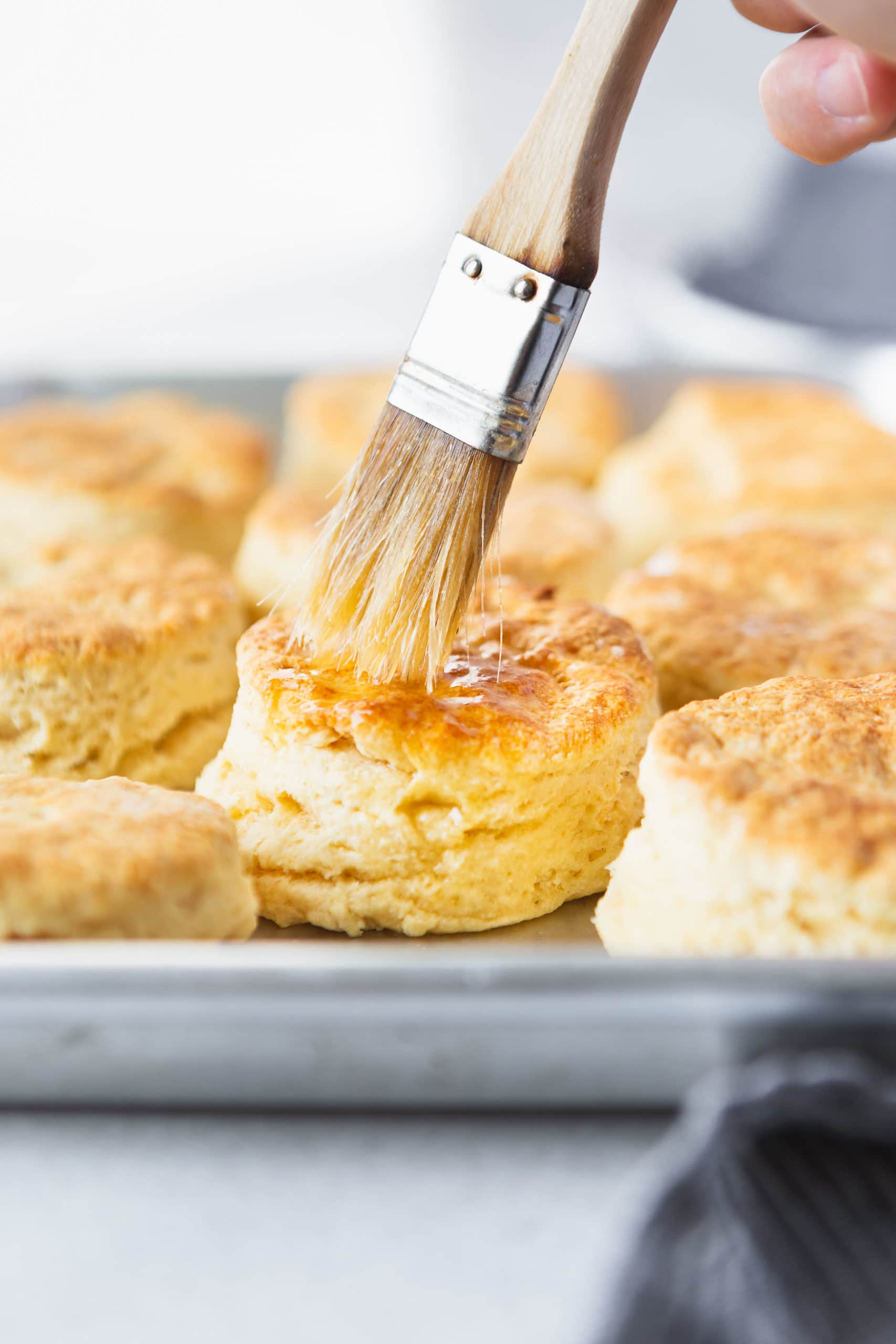 A pan full of golden brown buttermilk biscuits. Butter is being brushed on the top of a biscuit with a pastry brush.