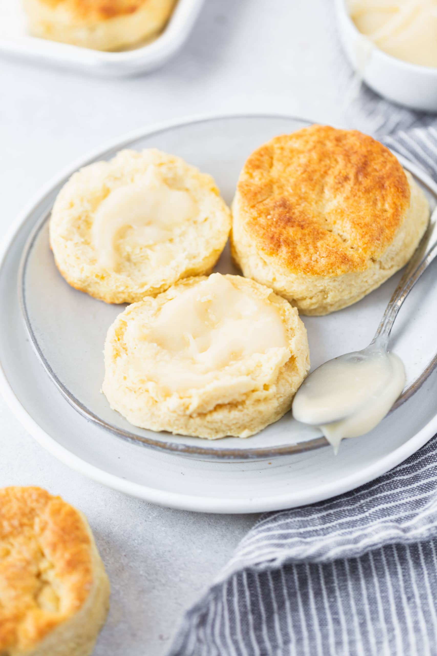 Two buttermilk biscuits on a small white plate. One biscuit is broken in half and has been buttered.