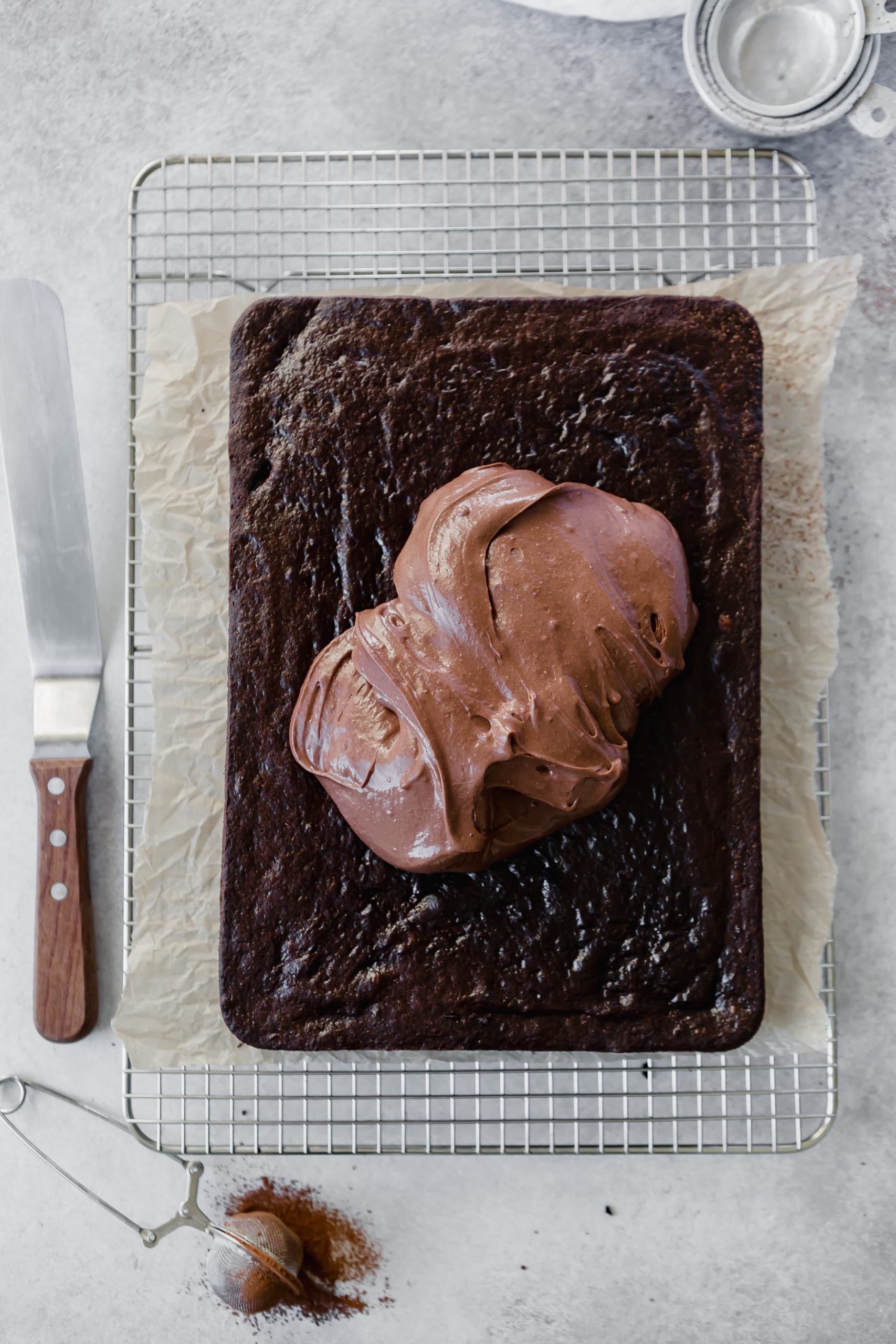 A 9x13 chocolate mayonnaise cake. There is a mound of chocolate frosting in the middle that has not been spread over the cake yet. A frosting spatula is next to the cake.