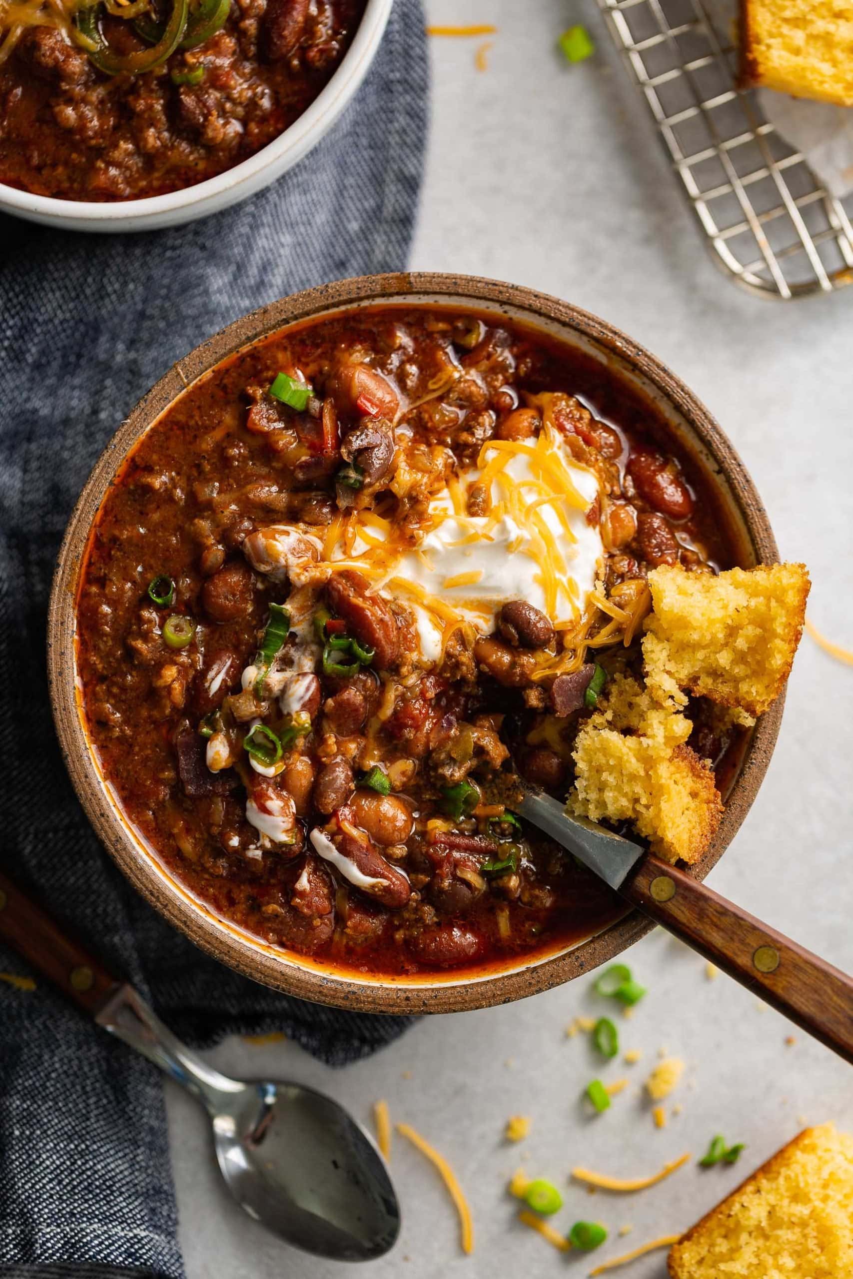 A bowl of the best chili. A spoon is in the bowl. There are beans and ground beef in a rich sauce and the chili is topped with sour cream and shredded cheese.