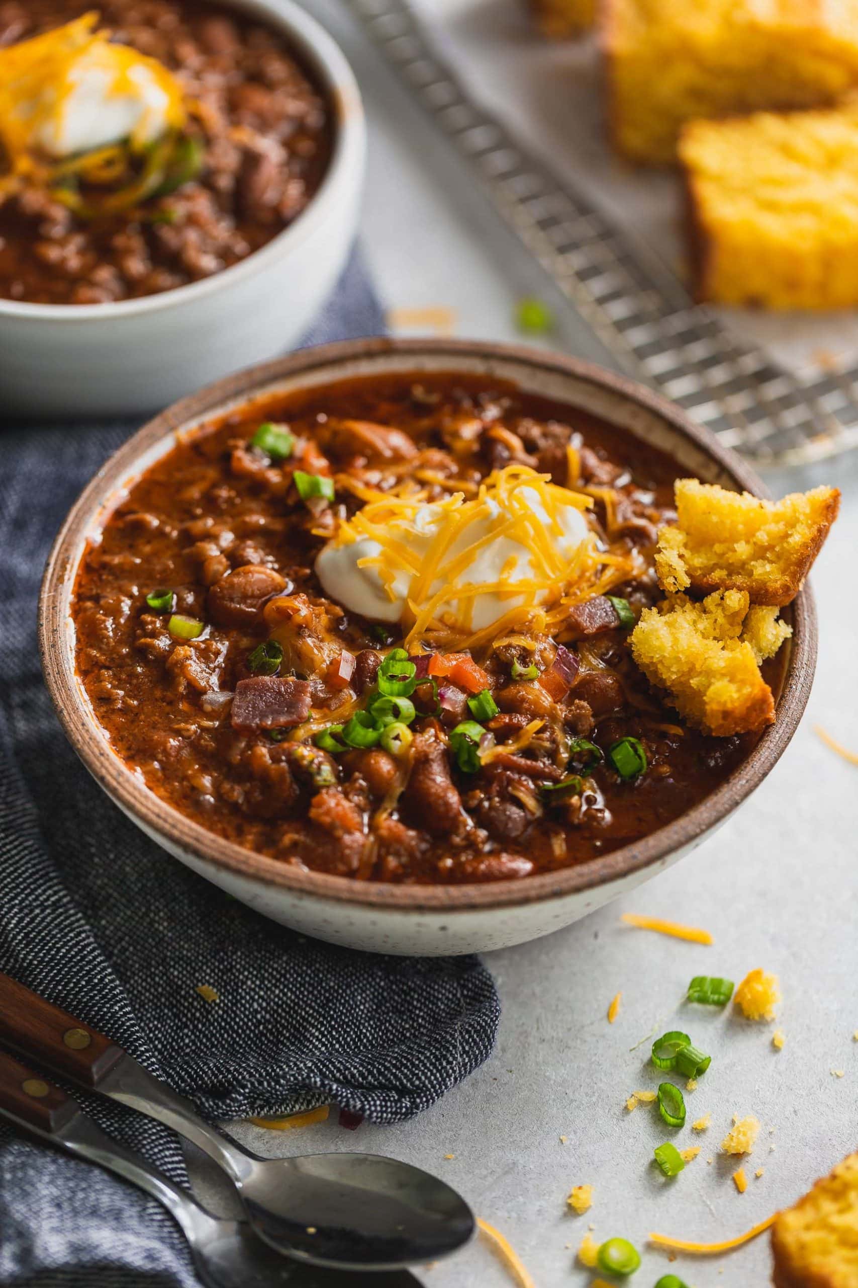 A bowl of the best chili. It has a spoonful of sour cream and shredded cheese on top. You can see the ground beef and beans in the chili, and a piece of cornbread is on the edge of the bowl. Another bowl of chili is in the background.